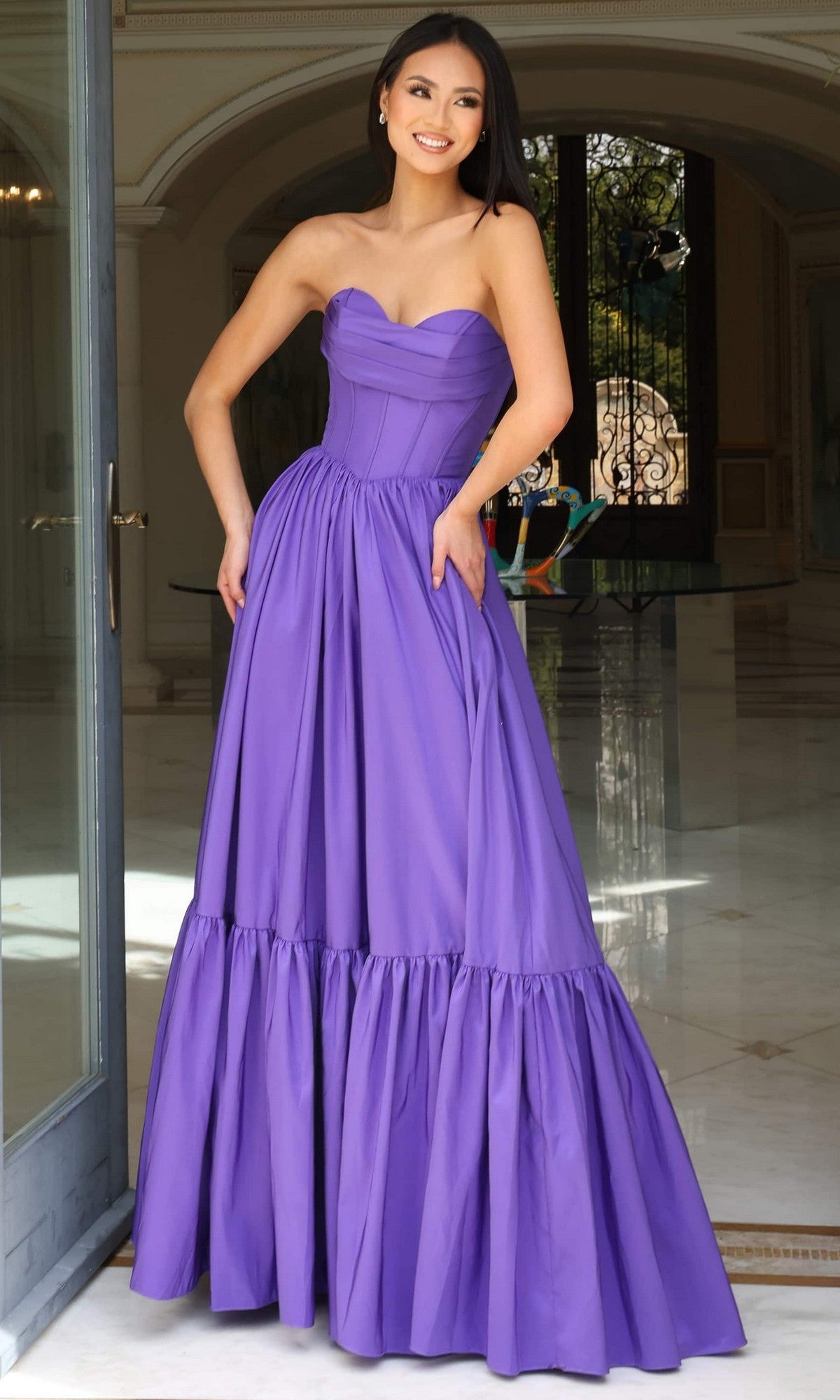A woman stands indoors in the Jovani 47883, a strapless long ruffled prom dress with a corset-style bodice and full gathered skirt, exuding elegance in deep purple.